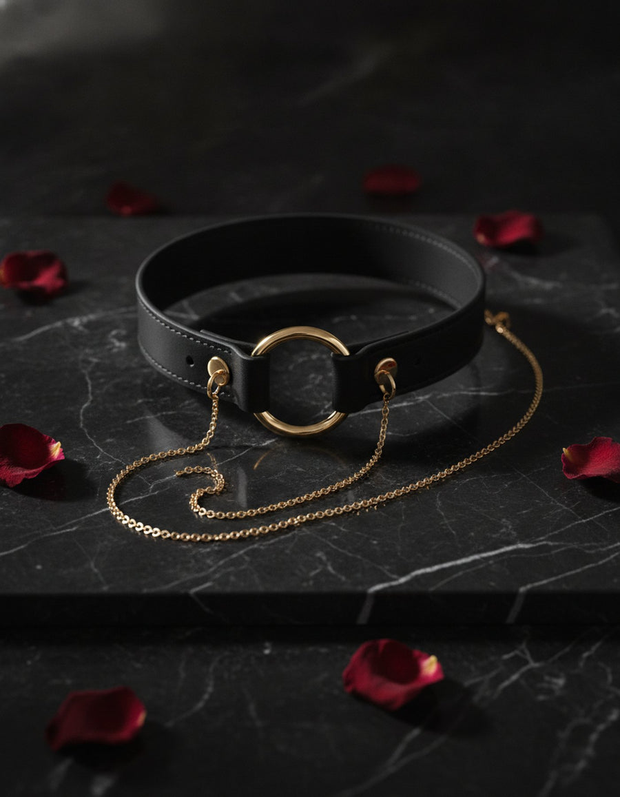 Black leather BDSM collar with gold O-ring and chain leash on marble